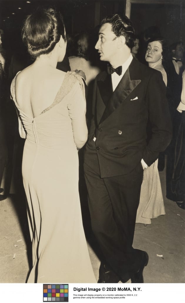 Archival photo of Margaret Scolari Barr and Salvador Dali at an exhibition opening. Barr is wearing a floor-length gown with her hair done up. Dali is wearing a tuxedo. Barr's back is to the camera while Dali looks off in the distance.