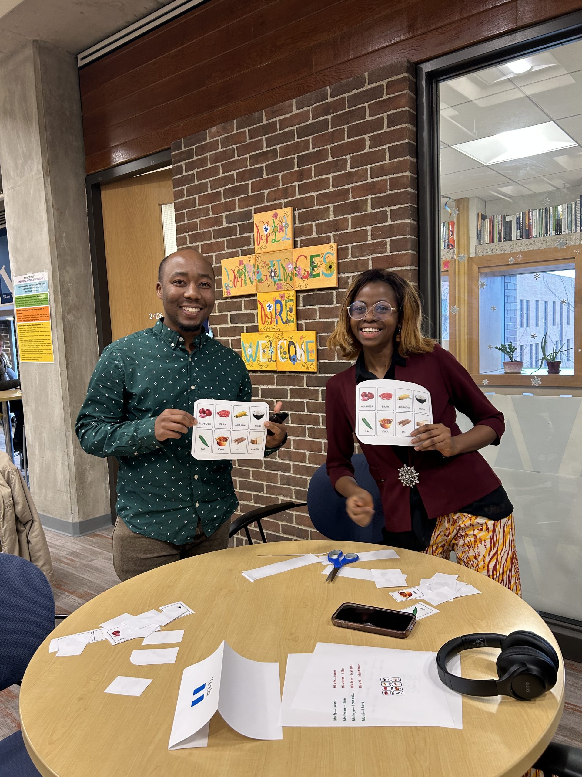 Two individuals standing, one teaching and another learning, both holding papers that share terms in Yoruba. In the background is a painted sign depicting, "all languages are welcome."
