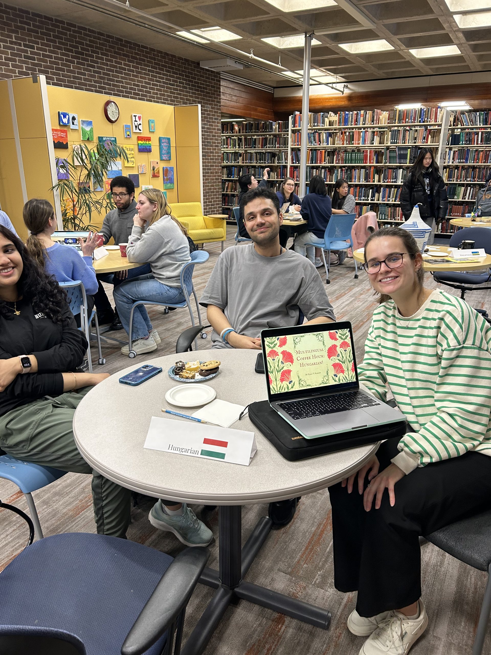 Table of three smiling people, one teaching Hungarian, the other two learning. The instructor uses her laptop which shows "Multilingual Coffee Hour, Hungarian" at a table with a sign reading, "Hungarian." In the background, there are tables of other people learning French and Karen.